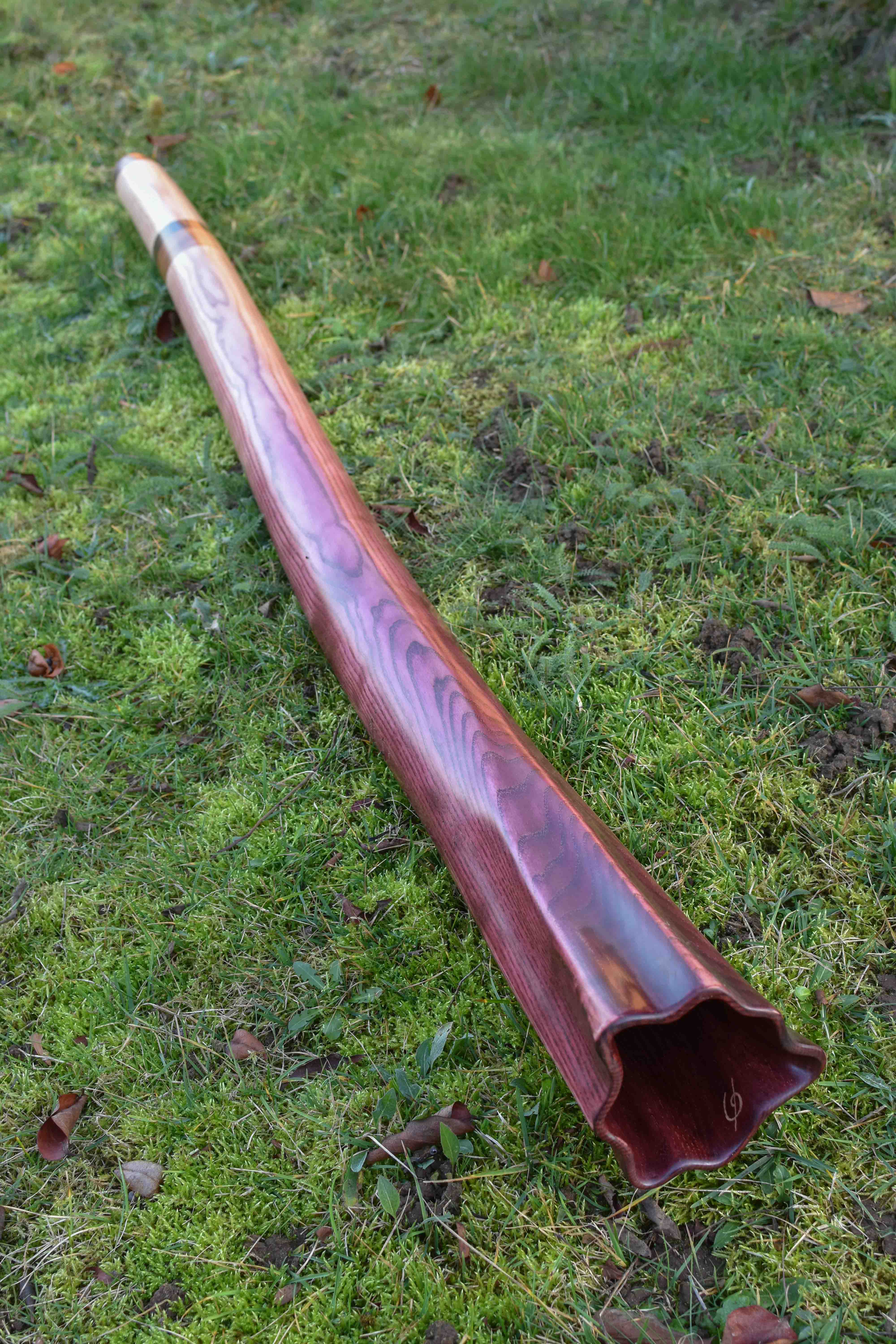 Didgeridoo nommé okumé, en Do, légèrement courbé, fabriqué dans un morceau de frêne, teinté en dégradé de pourpre, avec une cloche sculptée